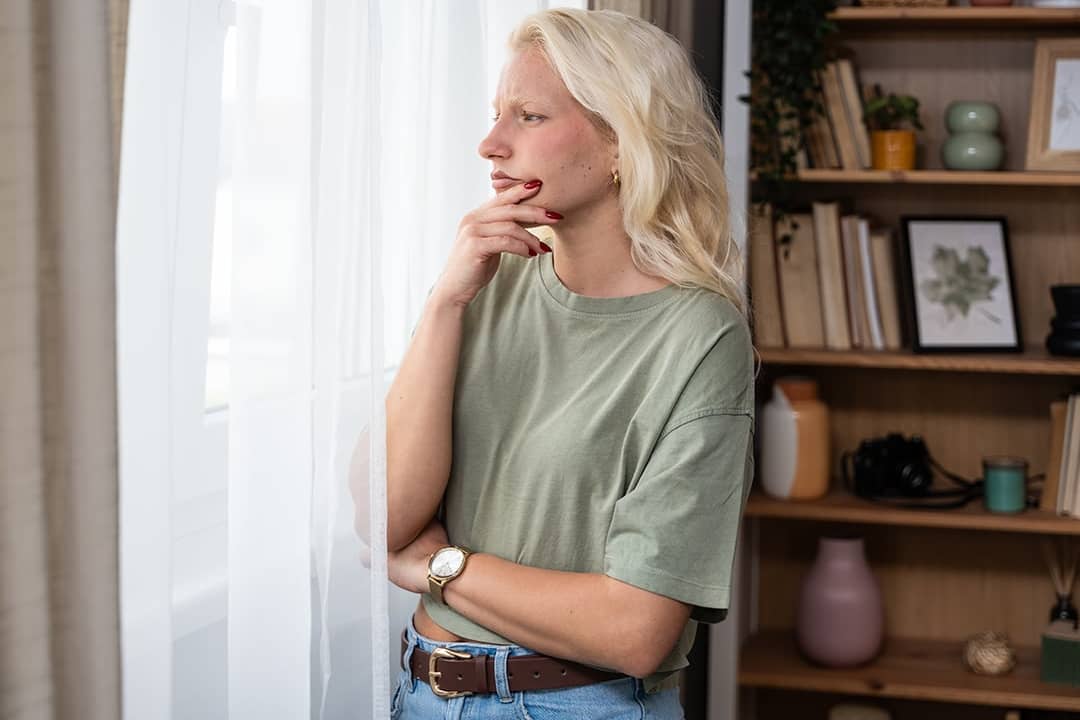A pensive young woman at home looking through her window.